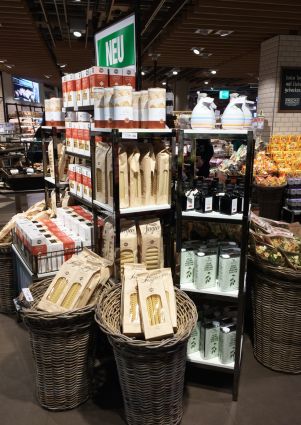 Italian food display at Migros. Pasta, olive oil, and other Italian products are on shelves and in baskets. Neu sign.