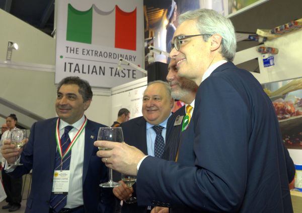 Italian producers toast resilience. Men with wine glasses at Italian food expo. The Extraordinary Italian Taste banner.