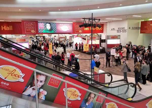Attendees at the Winter Fancy Food Show. Escalator with Specialty Food Association ads. Hall entrance and show signage visible.