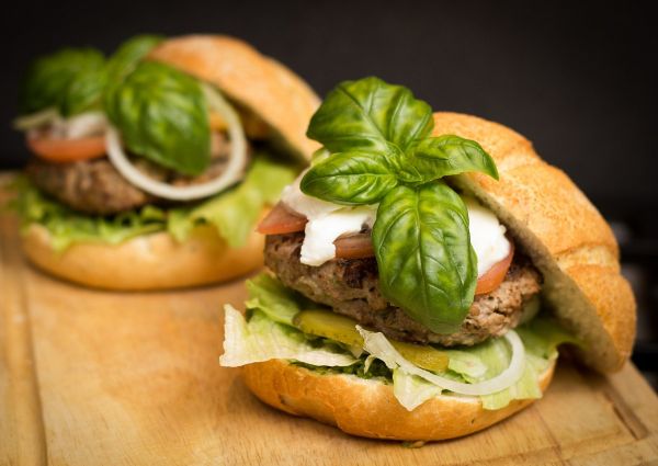 Two delicious hamburgers with basil, tomato, lettuce, onion, and cheese on a wooden board. Celebrate Hamburger Day!