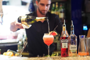 Bartender pours champagne into cocktail. Summer Resto Week drink special. Campari and El Jimador bottles.